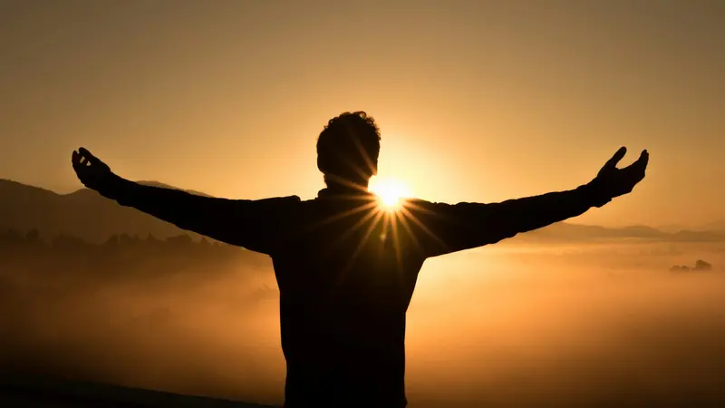 Person standing with arms open toward a warm golden sunrise
