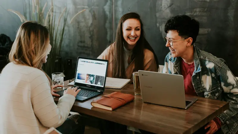 Three friends smiling and talking around a table with laptops